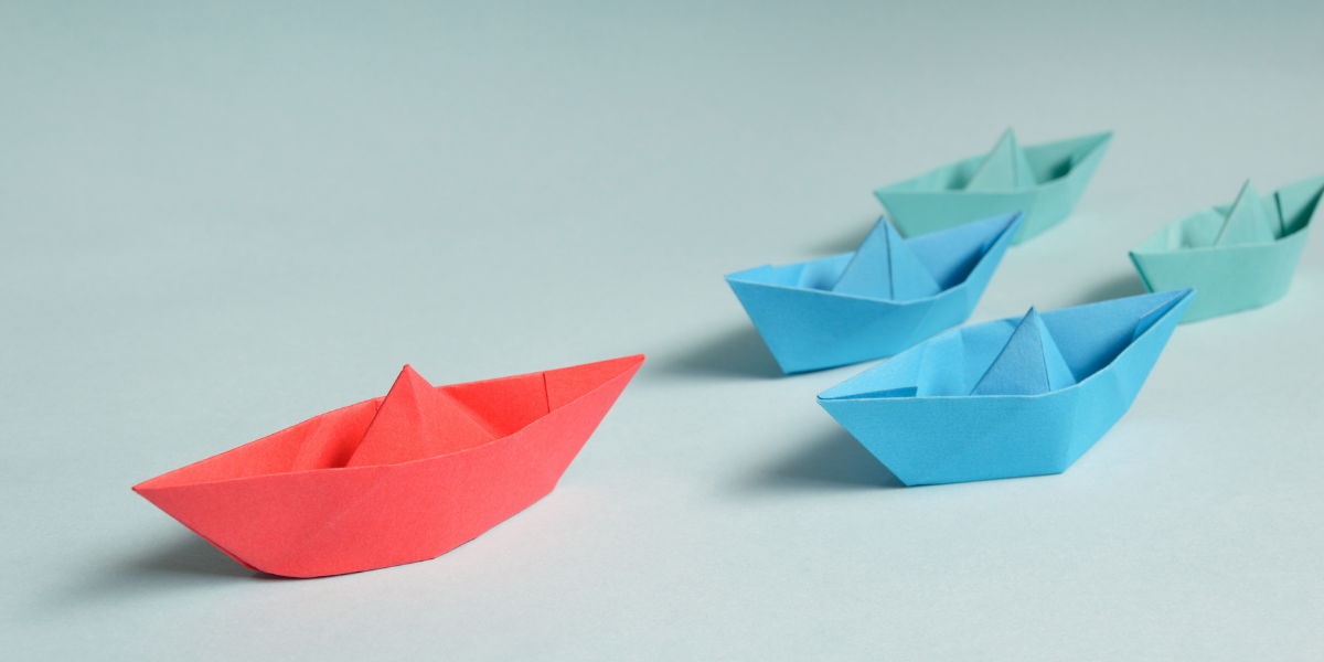 Five paper boats racing, with the red boat in the lead position. (All other boats are blue).