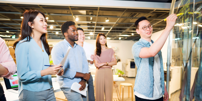 A group of people standing in an office watching someone present and write on the whiteboard.