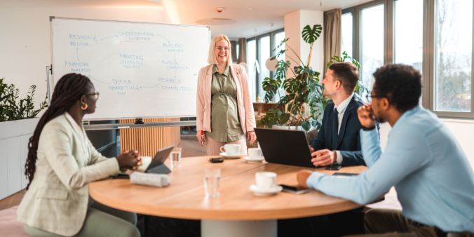 Three individuals are sat at the boardroom table and a woman is stood in front of a whiteboard presenting.