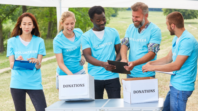 A group of volunteers in matching light blue shirts are standing under a white tent taking donations.