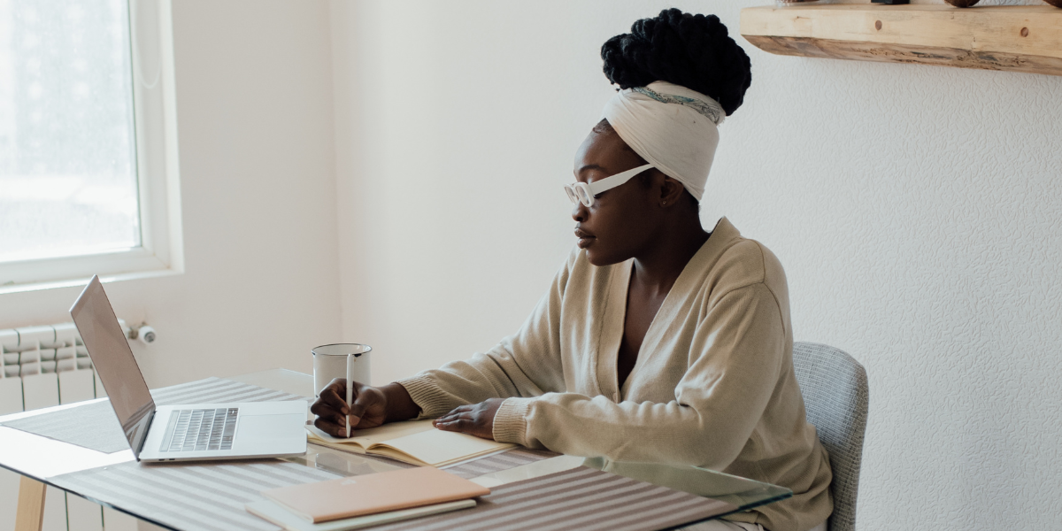 A woman is sat at a desk writing on a notepad with a laptop in front.