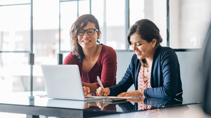 Two women are sat at a desk - one woman is writing in a notebook and the other is typing on a laptop.