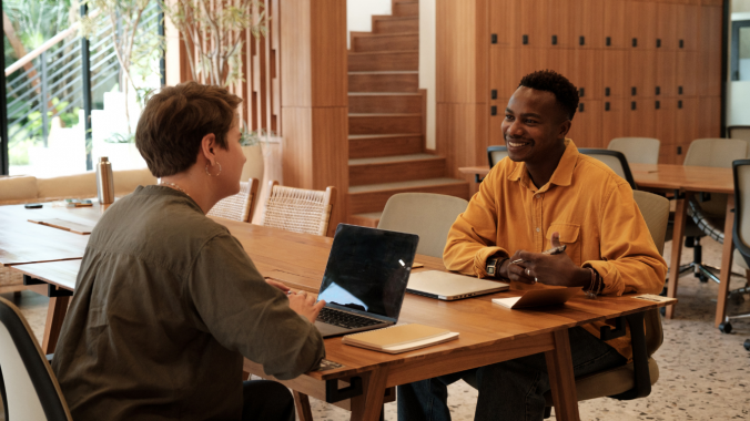 Two individuals are sat across from one another at a desk. The one individual is typing on a laptop.