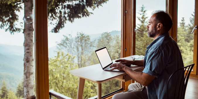 A man is sat in front of a wall of windows at a desk typing on a laptop.