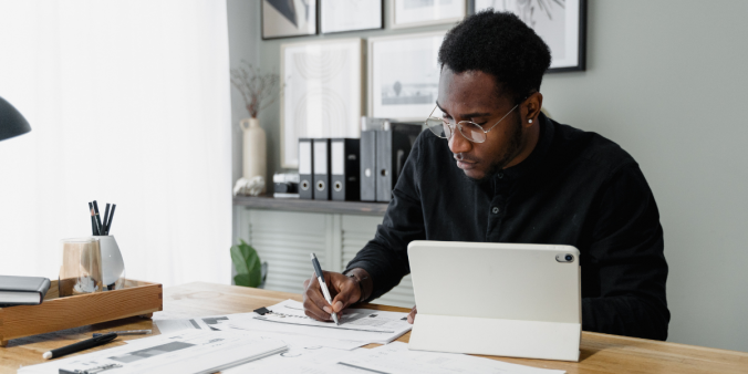 A man is sat at a desk writing on a piece of paper, with a laptop to the side.