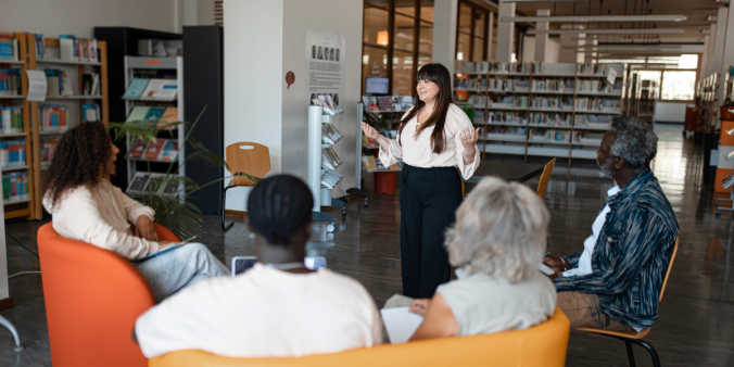 A group of individuals are sat in a half circle in a library. A woman is stood at the front of the room presenting.