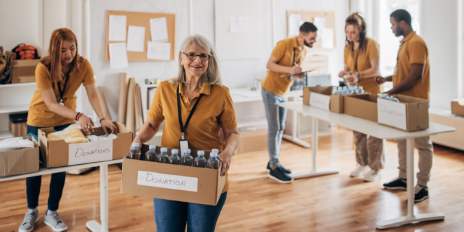 A group of individuals are volunteering at a food bank. They are all wearing matching yellow shirts. A woman is stood facing the camera holding a box of water bottles.