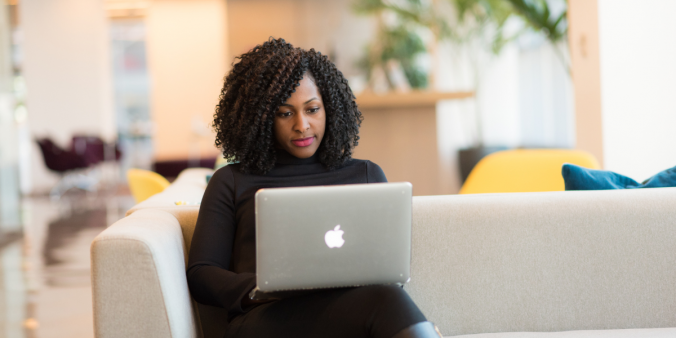 A woman wearing all black is sat on a beige couch typing on a laptop.