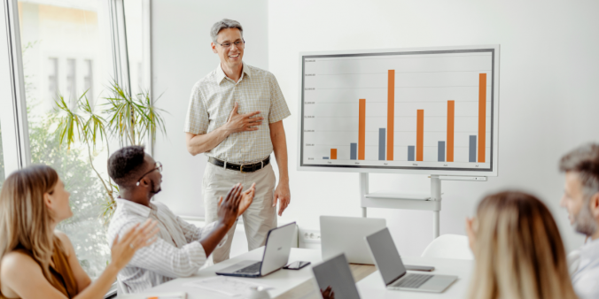 A man is stood at the front of a boardroom next to a whiteboard featuring statistics. The team members sat at the boardroom table are clapping.