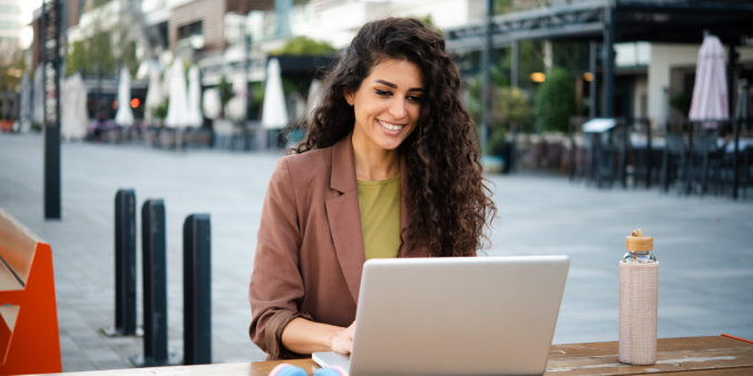 A woman is sat outside at a cafe typing on her laptop.