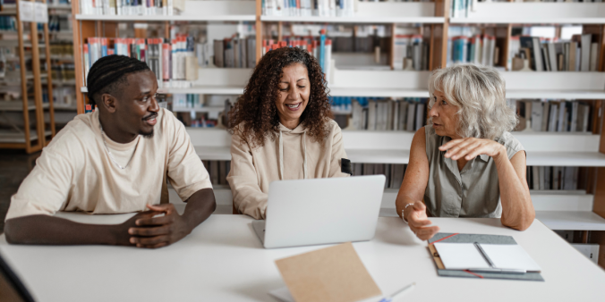 Three people are sat at a desk in a library. One woman is typing as they all chat.