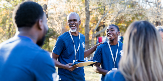 A group of volunteers in matching blue shirts are stood in a circle facing the coordinator holding a clipboard.