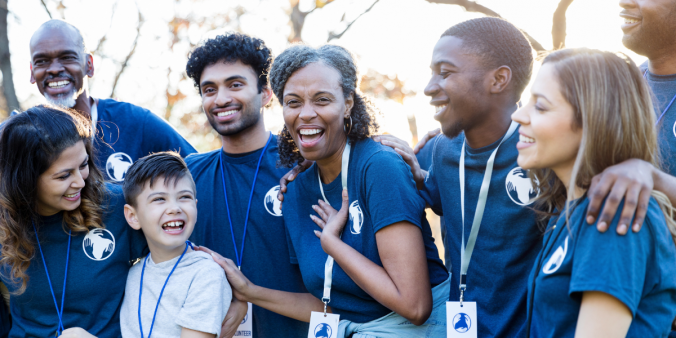 A group of volunteers is gathered standing arm in arm outside in the sunlight. They are all wearing matching blue volunteering shirts and appear to be laughing.