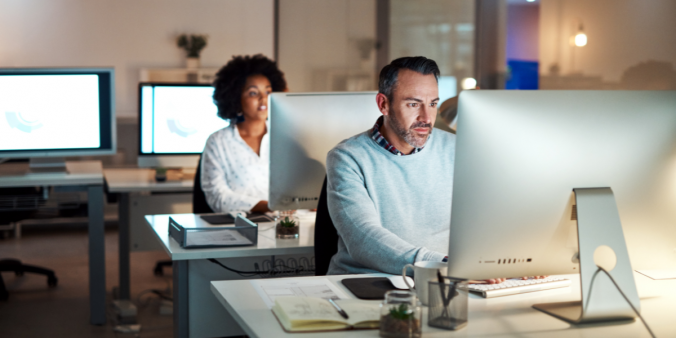 Two people are sat in an office at their desks, it appears to be the later evening.