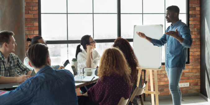 A group of people are sat at a boardroom table. One man is standing presenting to the group.