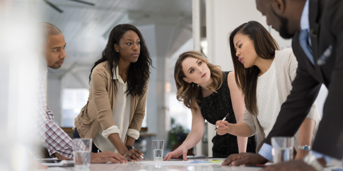 A group of individuals are stood around a boardroom table working on a project.