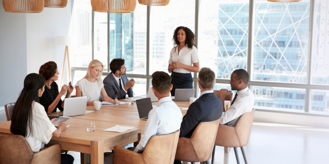 A woman is stood at the head of a boardroom table in front of a wall of windows presenting to a group of individuals.