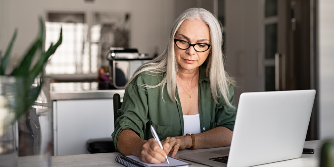 An older woman wearing a green shirt is sat at a white desk writing on a notepad.