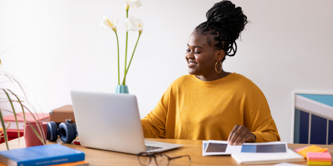 A woman in a yellow shirt is sitting in a well-lit room. The woman is sat at a desk typing on a laptop.