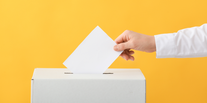 An individual placing a white piece of paper into a white ballot box. The background of the image is yellow.