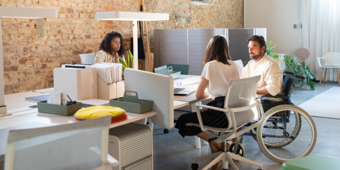 Two women are sat at a desk working, and one man in a wheelchair is talking with one of the women.