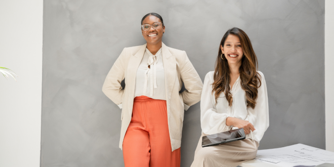 One woman is standing and one woman is sitting holding a tablet. They are both looking at the camera and smiling.