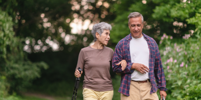 Two older individuals are walking outside in nature arm in arm chatting.