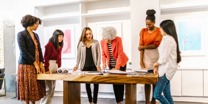 A group of individuals are stood at a boardroom table. One of the women is pointing at a document on the table.