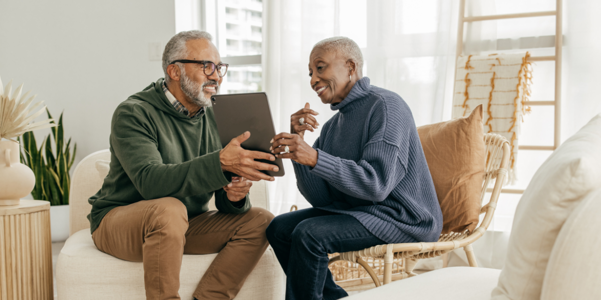 Two older individuals are sat across from one another. The older man is holding a tablet up showing something to the woman.