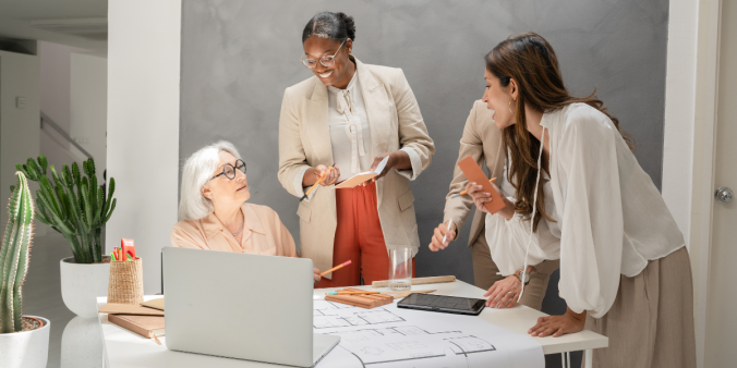 A group of women are standing at a boardroom table. One woman is sitting with a laptop in front of her. They all appear to be collaborating.