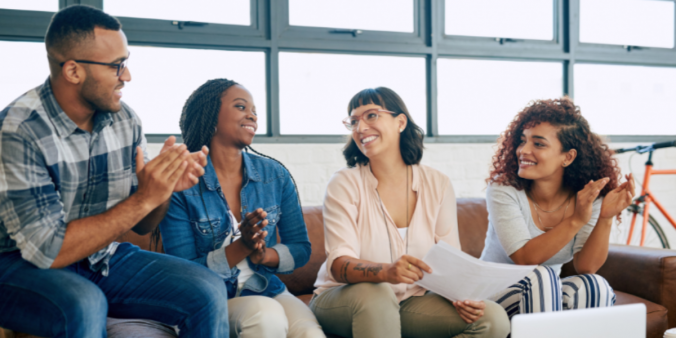 Four people sitting on a couch conversing appearing to have a meeting. Three of the individuals are clapping appearing to celebrate the one individual.