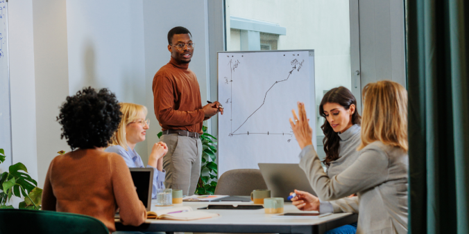 Four women are sat at a boardroom table. Two of the women are typing on their laptops. One man stands at a chart at the front of the room presenting.