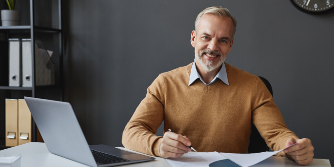 An older man is sitting at a desk writing on a piece of paper and smiling at the camera.