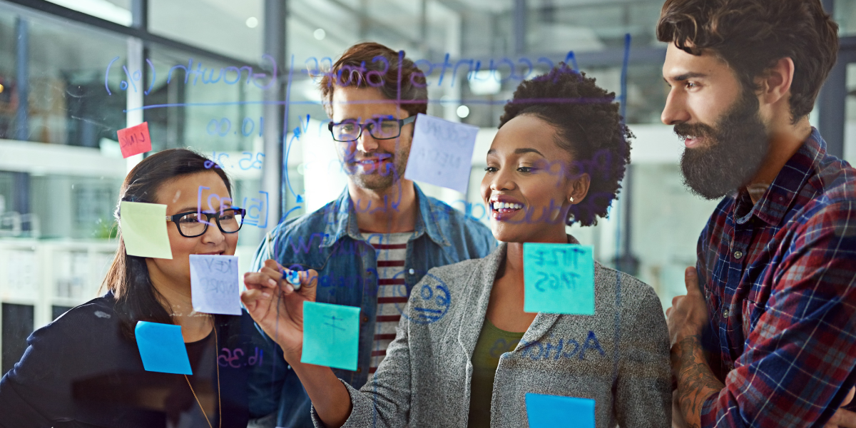 A group of four individuals are facing a whiteboard and one woman is writing on the board as the others appear to be collaborating.