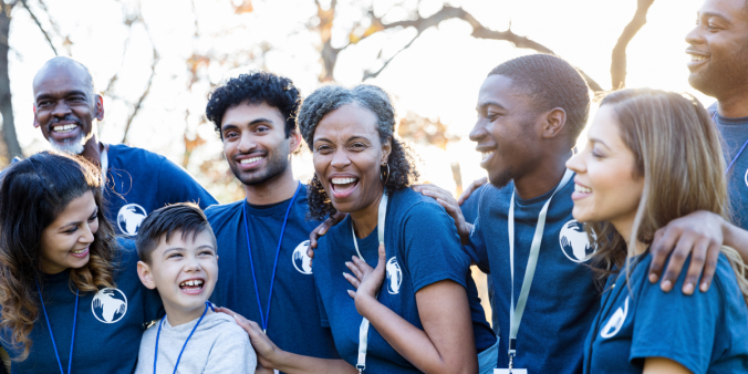 A group of volunteers is gathered standing arm in arm outside in the sunlight. They are all wearing matching blue volunteering shirts and appear to be laughing.