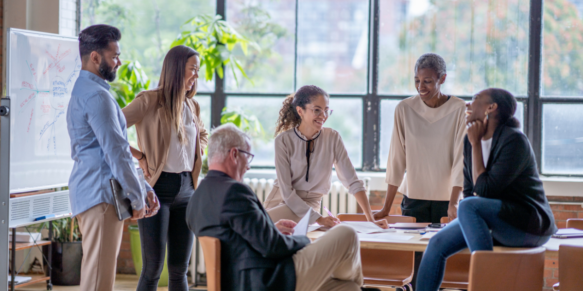 Six people sitting in a boardroom in front of a panel of windows talking.