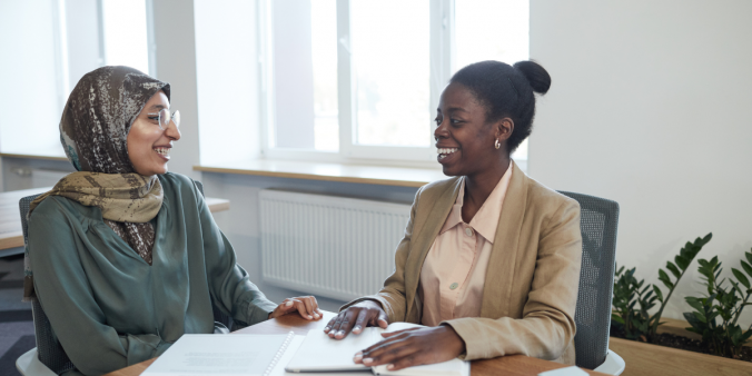 Two women sitting at a desk facing each other smiling and chatting.