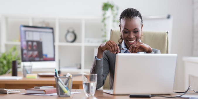 A woman sitting at a desk looking at her laptop and holding her glasses.