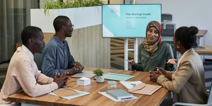 Four people sitting at a boardroom table conversing with a tv in the background displaying meeting notes.