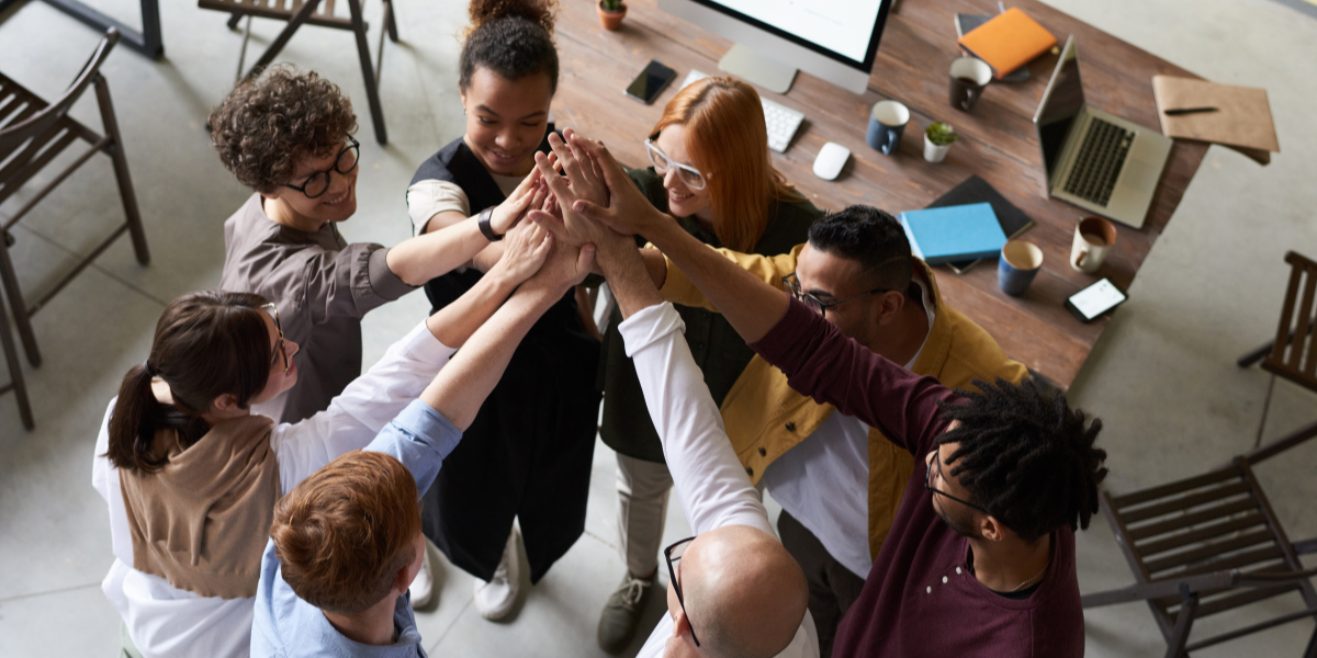 A group of people standing in a circle with their hands touching - signifying teamwork.