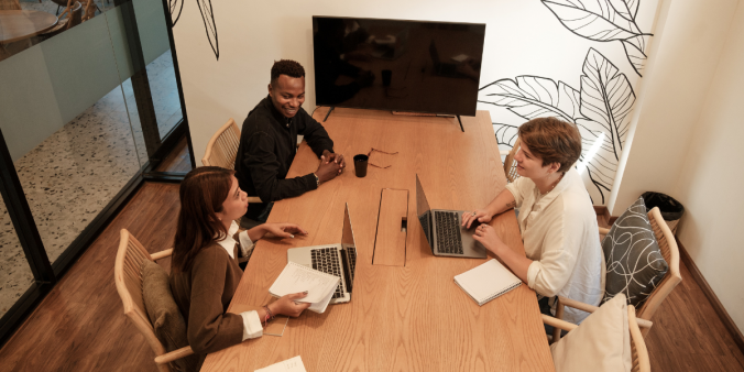 Three people sitting at a board room table conversing with their laptops on the desk.